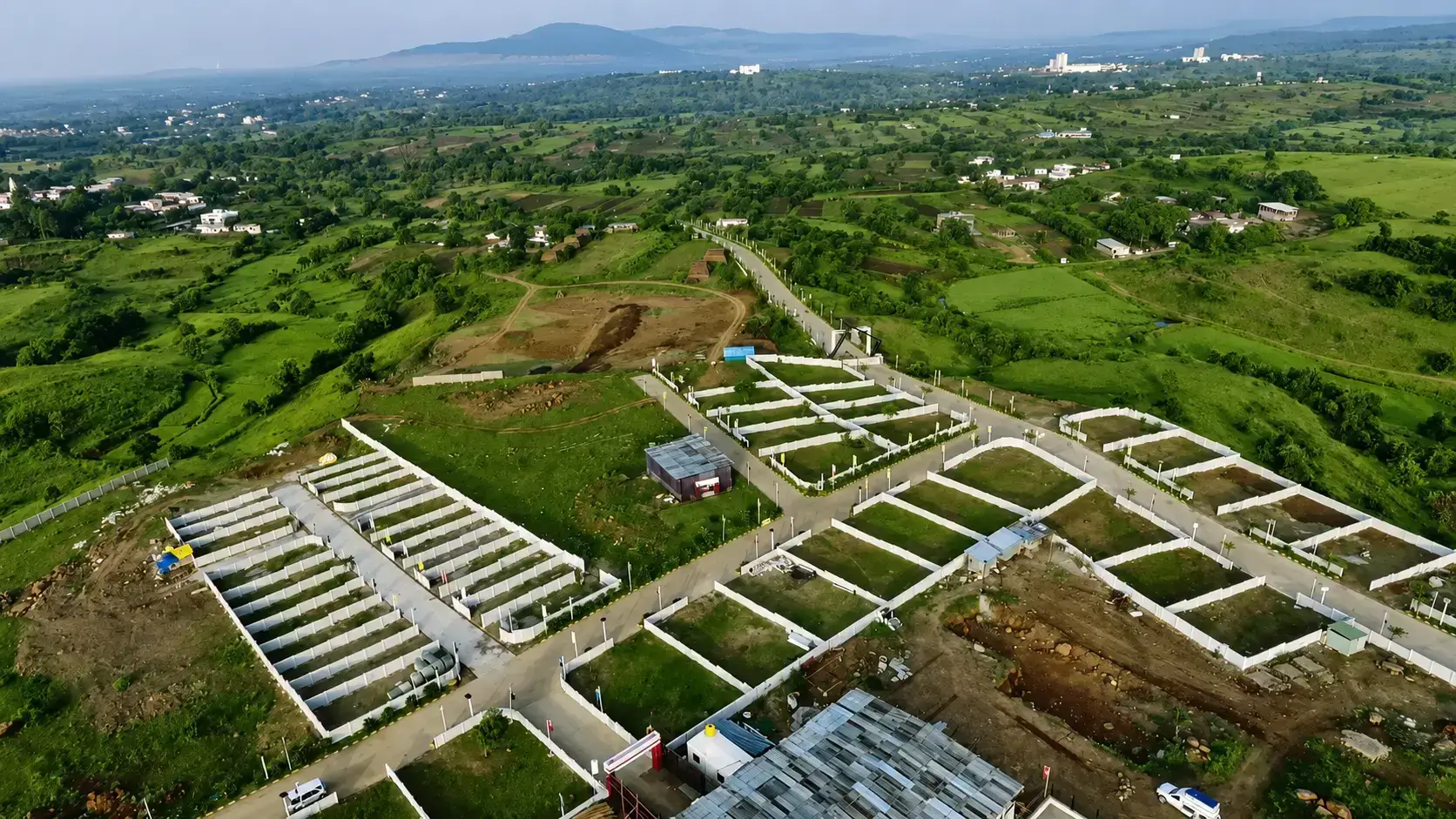 Aerial drone view of plotted development infrastructure and clear land parcels surrounded by a lush green hilly landscape in the Kikvi area.