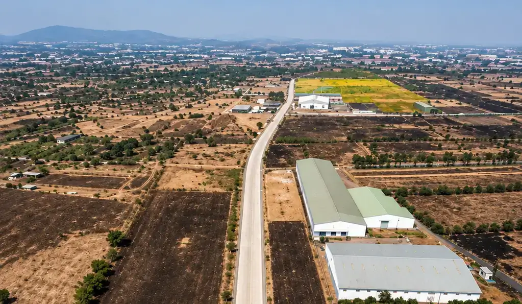 Aerial drone view of long warehousing sheds and a concrete access road situated amid open terrain and agricultural fields in the Nasrapur industrial area.