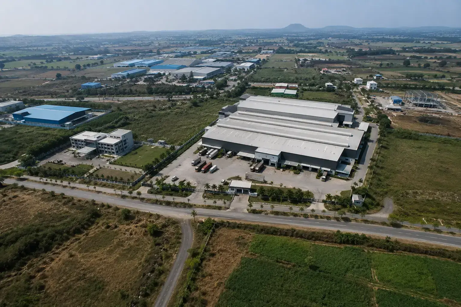 Aerial view of a large logistics warehouse and administrative building surrounded by open green landscape in the Phulgaon industrial area.