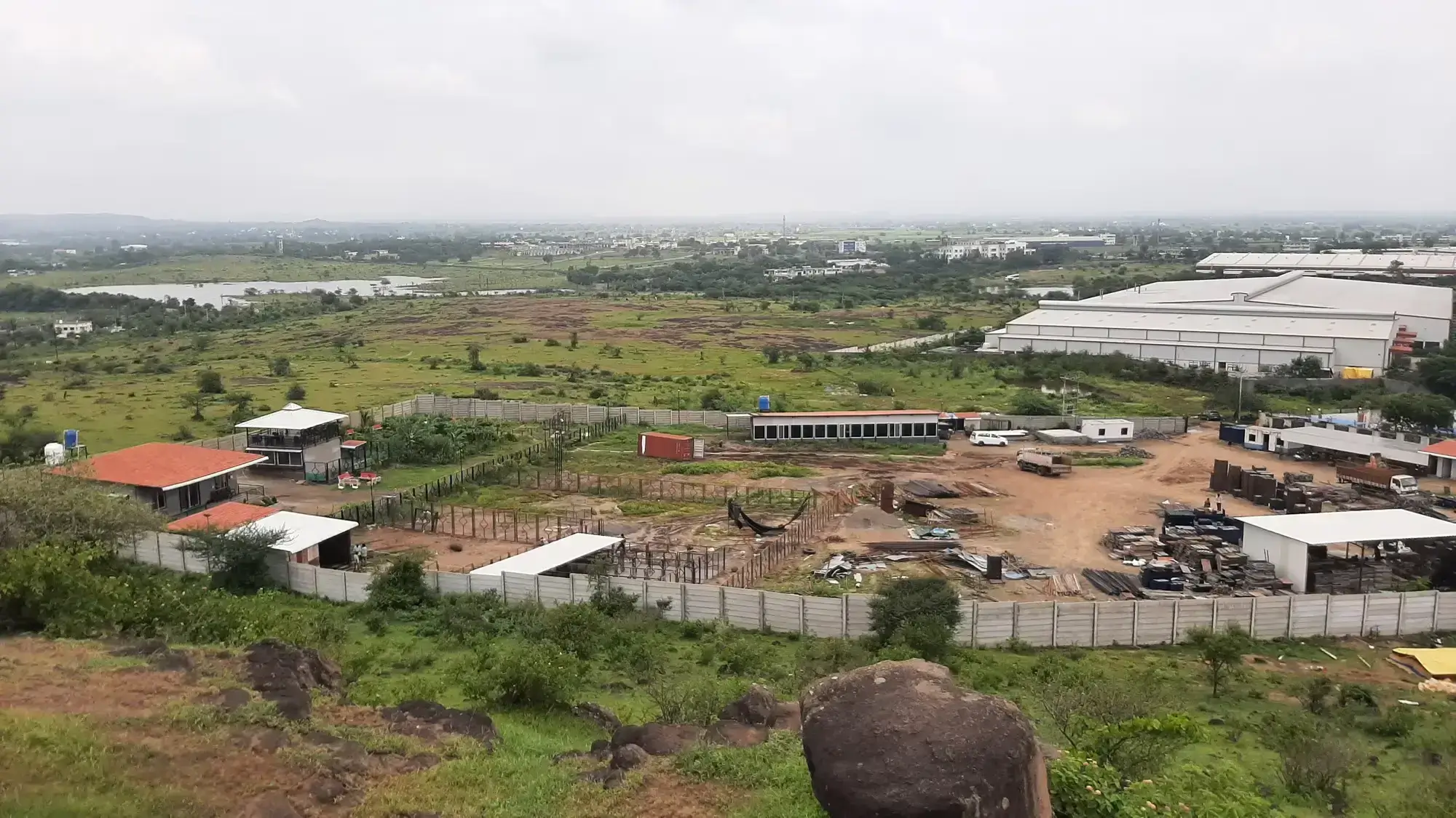 Elevated view of a fenced industrial construction site and surrounding green landscape in the Sanaswadi industrial area along the Pune-Nagar highway.
