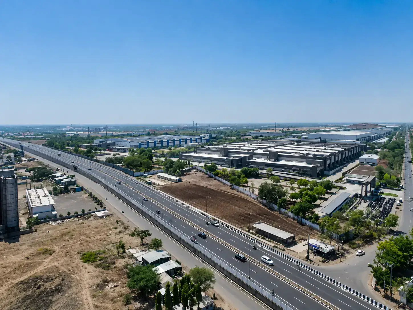 Aerial drone view of a multi-lane highway stretching alongside expansive industrial sheds, manufacturing plants, and green spaces in the Shirwal industrial area.