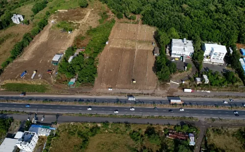 Aerial view of a major multi-lane highway running alongside cleared industrial plots and commercial buildings bordering a lush green landscape in the Khed Shivapur area.